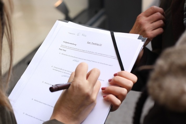 Eine Person mit langen Haaren mit Stift in der Hand f&uuml;llt ein Formular auf einem Klemmbrett aus- gegen&uuml;ber steht eine Person, die von ihr befragt wird