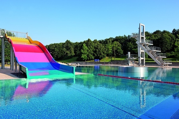 Ansicht Freibad mit farbiger Rutsche und Sprungturm im Hintergtund, dahinter große Bäume