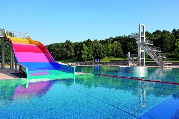 Ansicht Freibad mit farbiger Rutsche und Sprungturm im Hintergtund, dahinter gro&szlig;e B&auml;ume