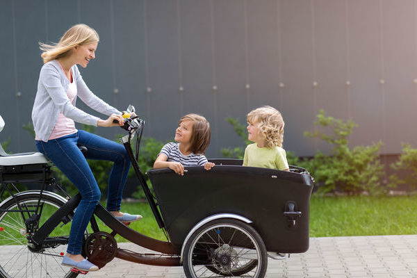 Eine Frau fährt auf einem Lastenrad. Im Lastenkorb sitzen zwei Kinder Eine Frau fährt auf einem Lastenrad. Im Lastenkorb sitzen zwei Kinder