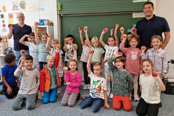 18 Kinder und zwei Erwachsene als Gruppe in einem Klassenzimmer. Jeder hält einen Apfel in der Hand. 
